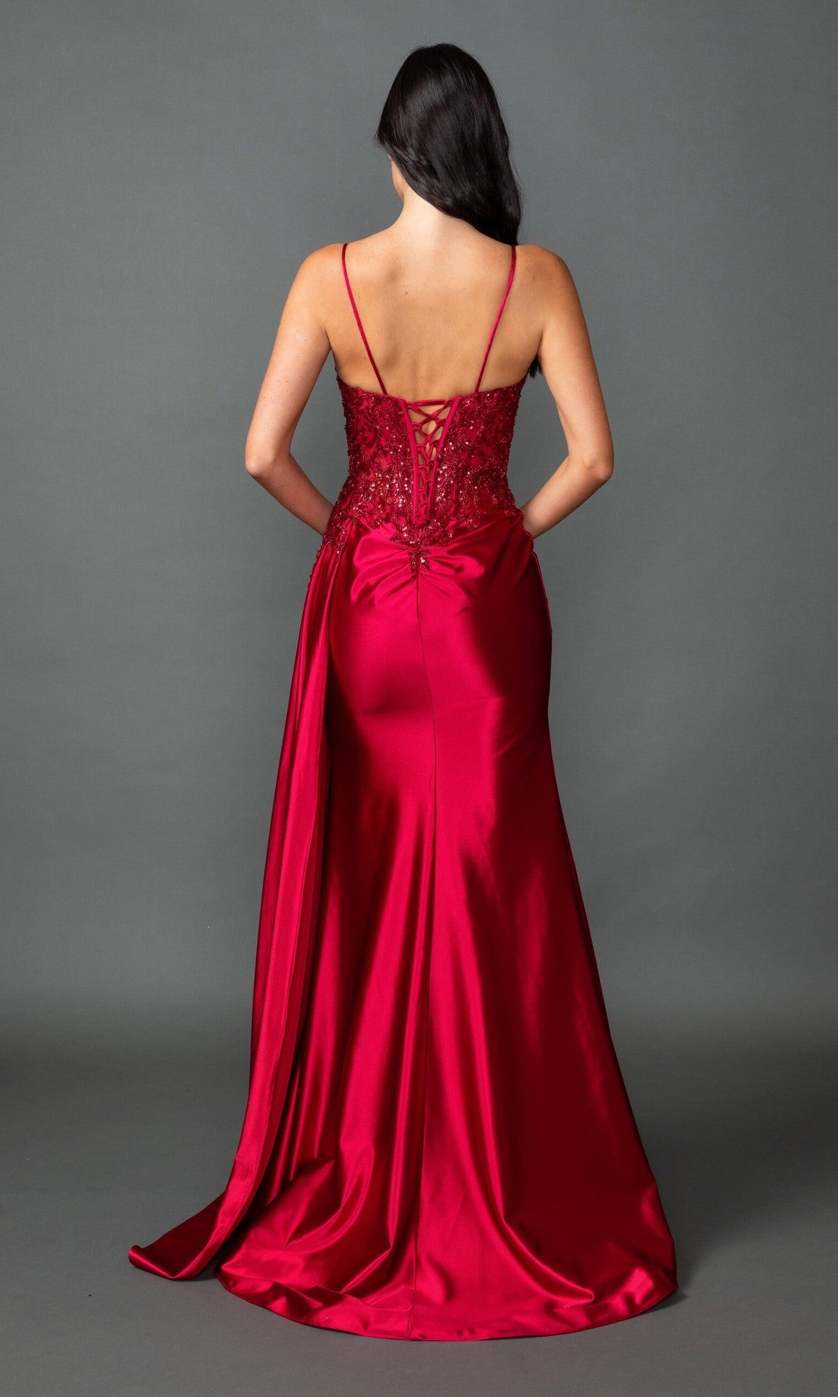 A woman faces away in the Long Prom Dress: Dancing Queen 4552, featuring red satin, a corset bodice, lace-up back, thin straps, and a floor-length skirt against a plain gray background.